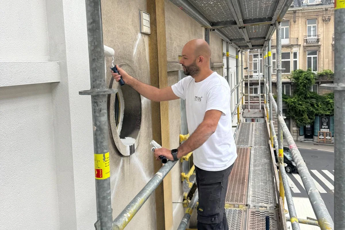Medewerker van MOS Reinigingsservice die vanaf een steiger met een rol verf aanbrengt rond een ovaal gevelraam; links is de crepi al wit afgewerkt.