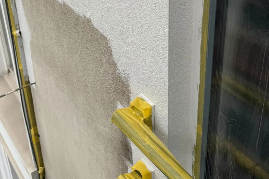 Gevel van dichtbij vanaf de steiger: ramen en kaders afgeplakt met geel tape, een vlak oude grijze crepi naast de eerste witte laag.