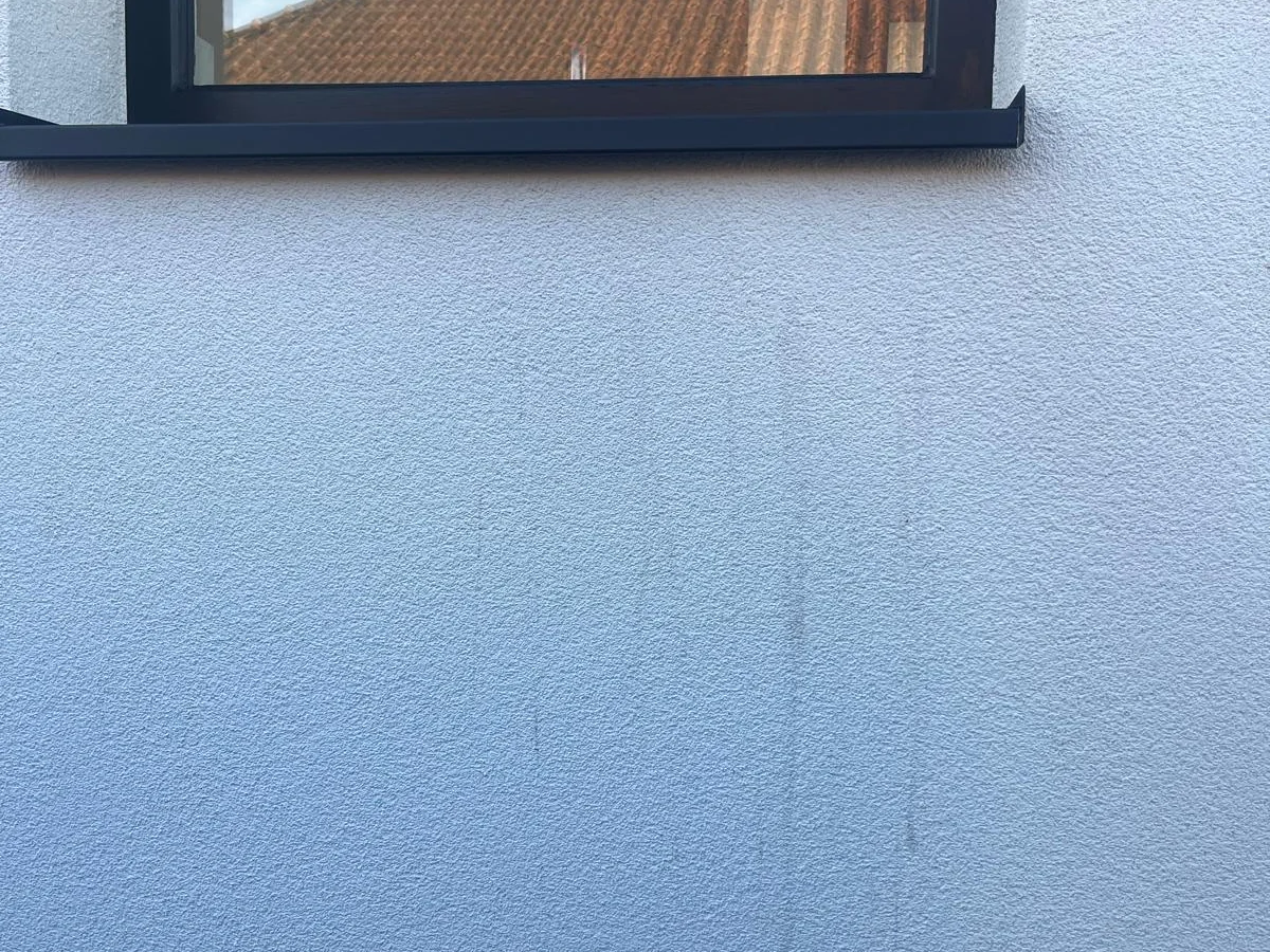 Witte crepigevel onder een donker raamkader, met lichte grijze strepen die van de dorpel naar beneden lopen.