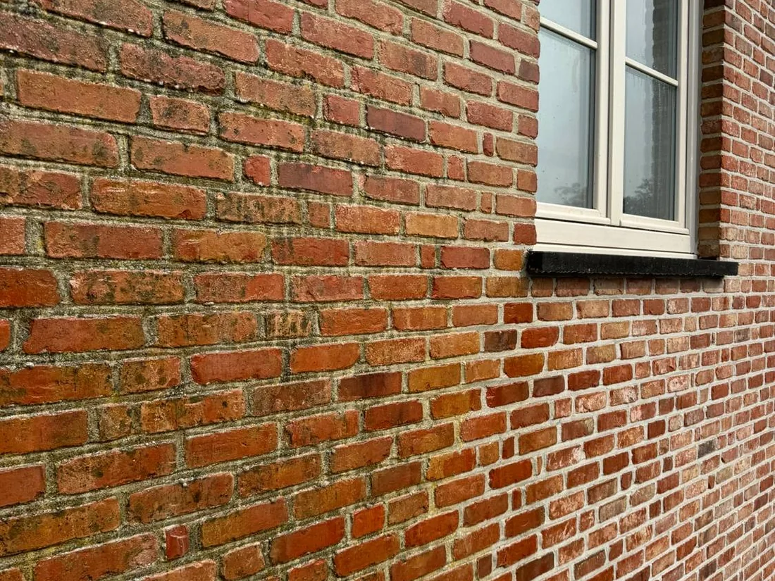 Bakstenen gevel van dichtbij: links vuile stenen en donkere voegen, rechts naast het raam gereinigd.